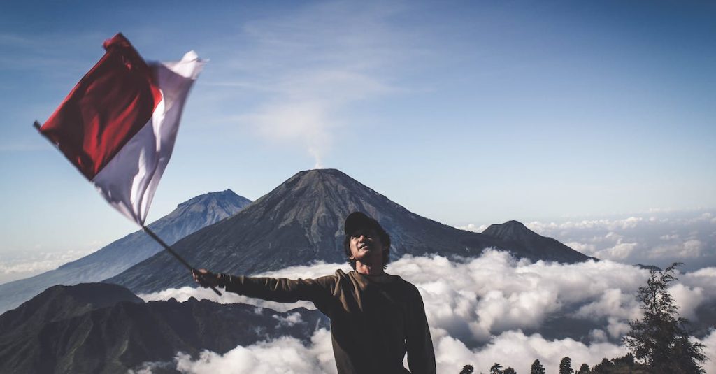 man-wearing-black-crew-neck-sweater-holding-white-and-red-flag-standing-near-mountain-under-blue-and